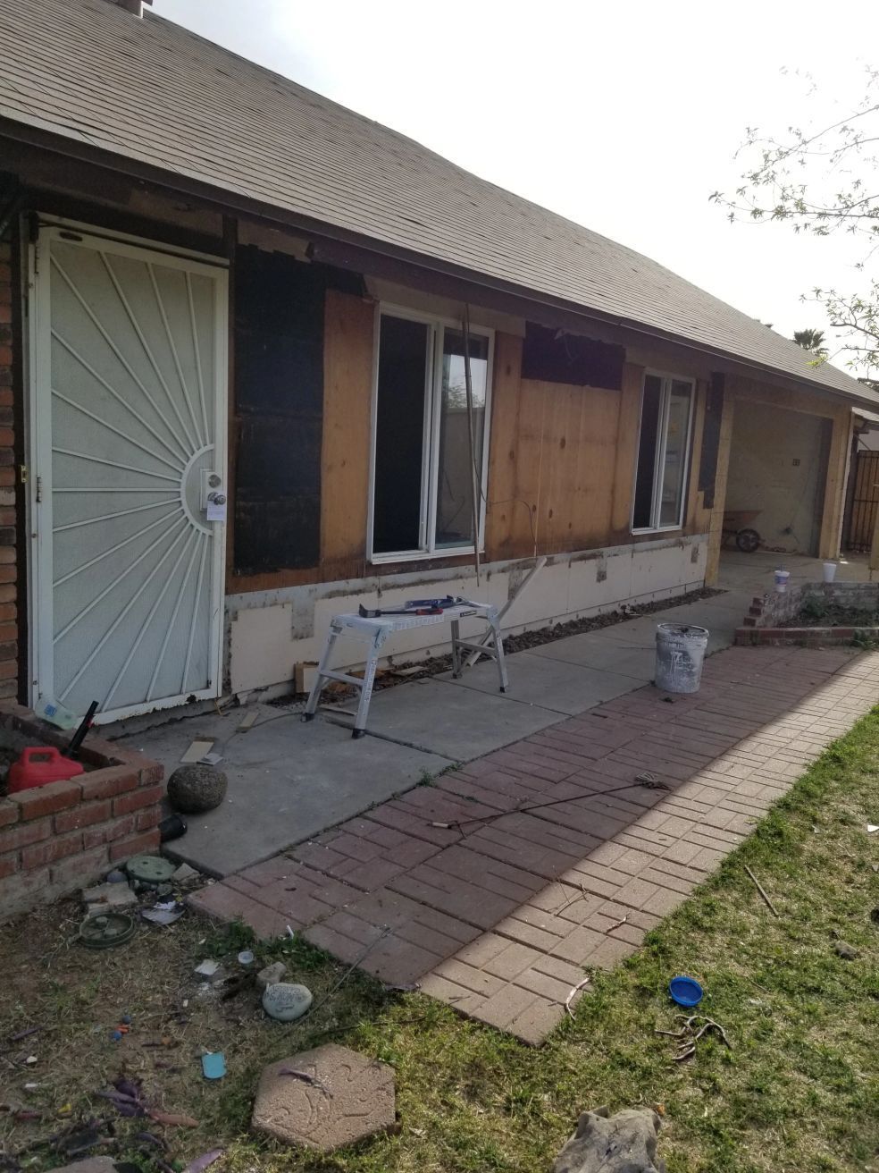 Rundown house exterior with boarded windows, white door, and a brick path along the side.