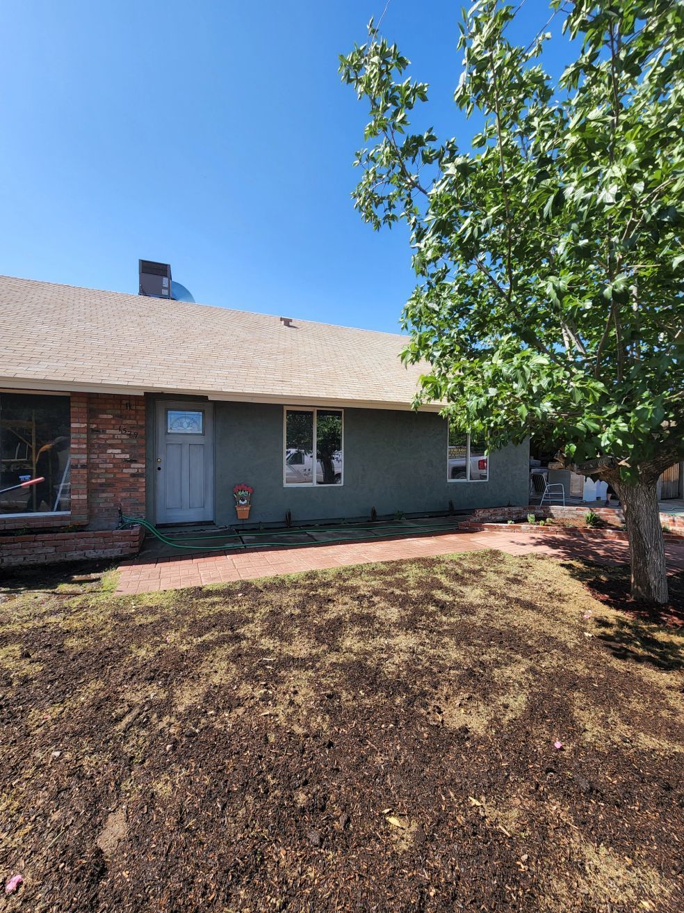 Single-story brick house with a gray roof, tree in front yard, and patchy brown lawn under a blue sky