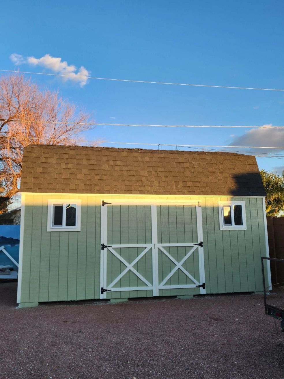 Small pale green shed with white trim, brown shingled roof, and double barn doors under a blue sky.