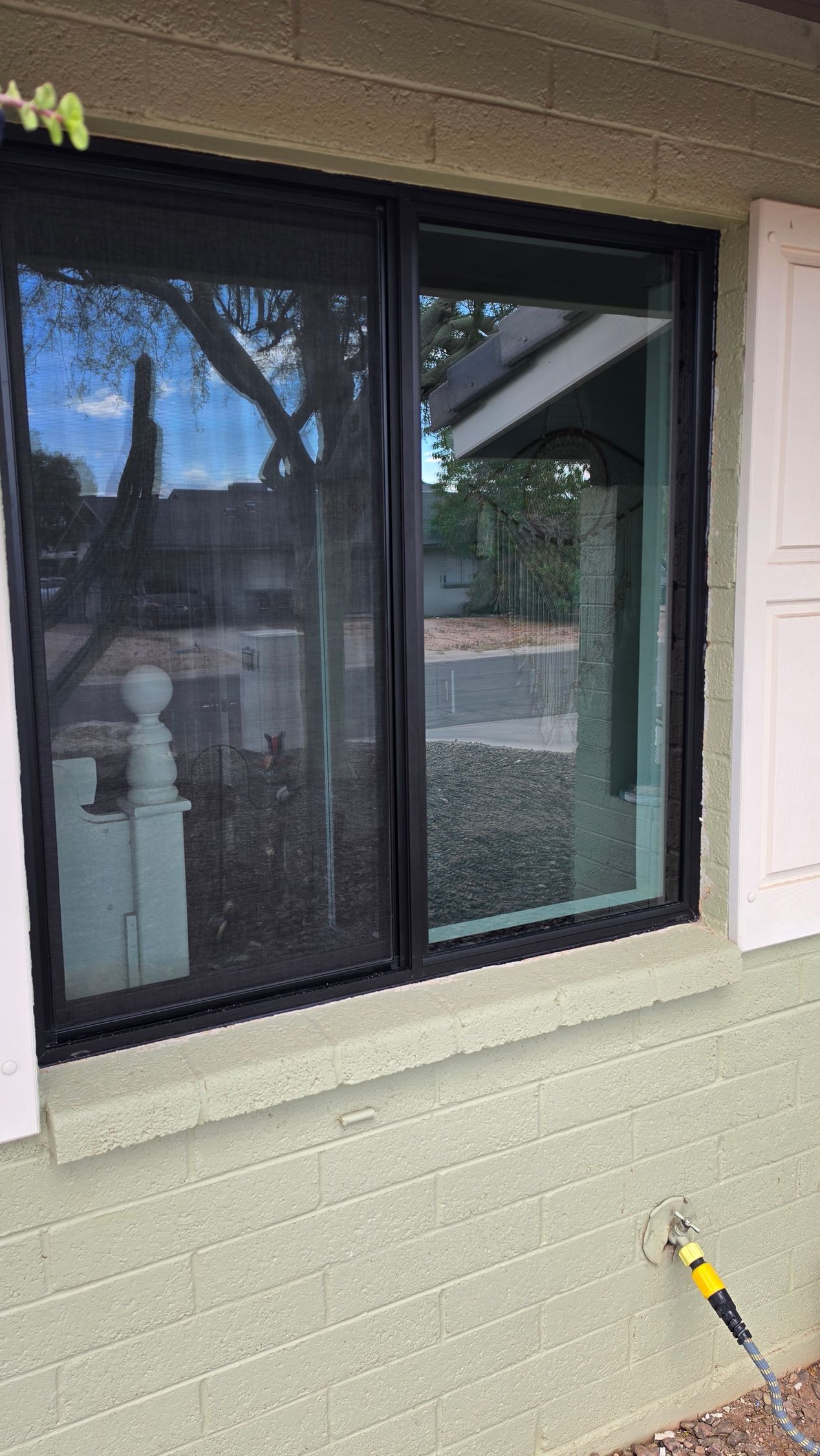 Black-framed window in green siding wall, reflecting trees and porch, with a yellow hose at bottom right