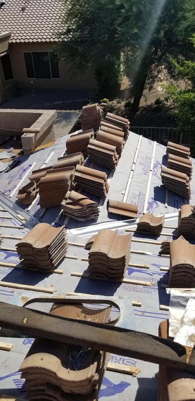 Stacks of tan roof tiles and white underlayment on a sloped roof under construction, with tools nearby.