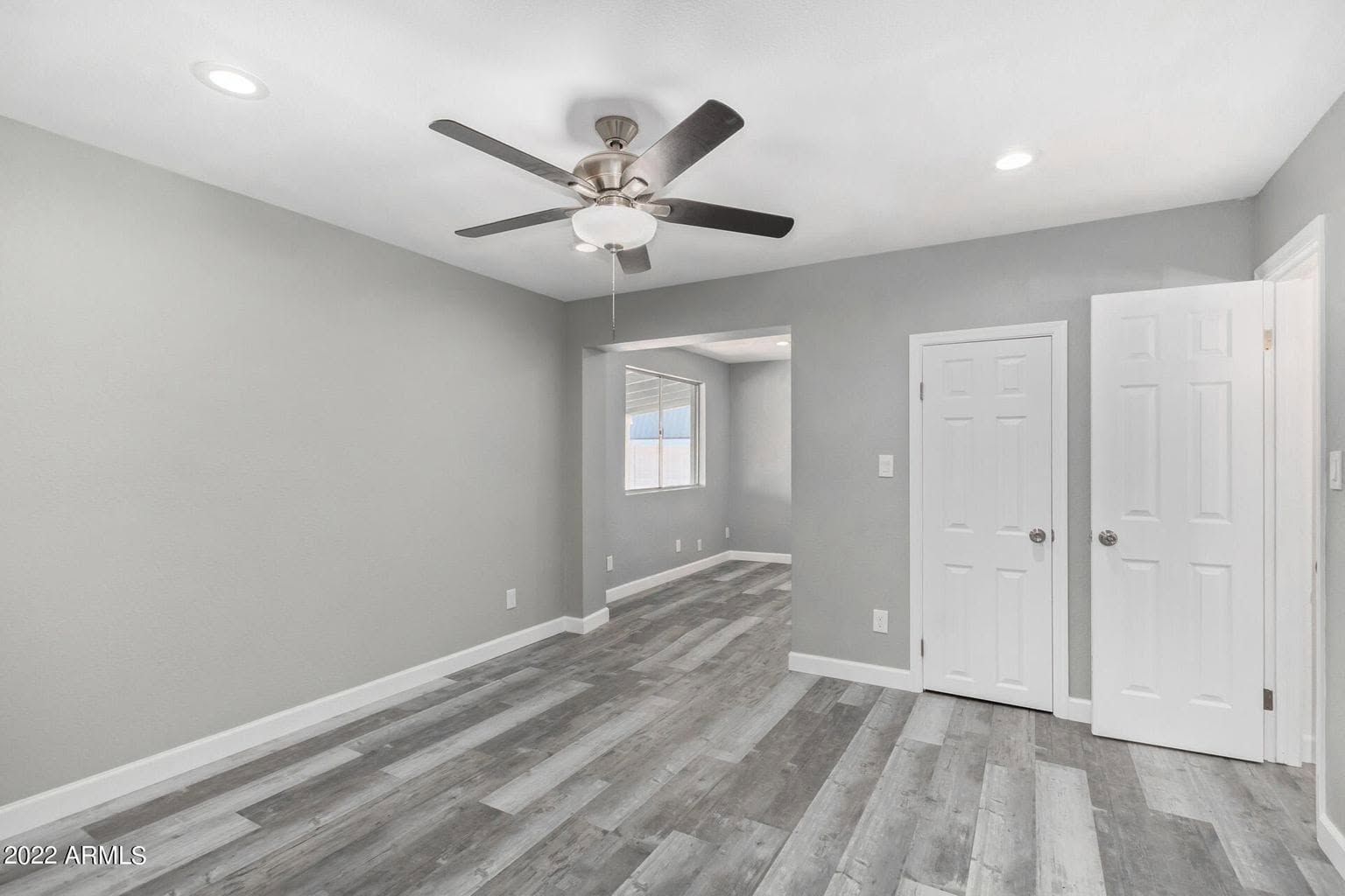 Empty gray bedroom with light wood floors, ceiling fan, and white closet doors