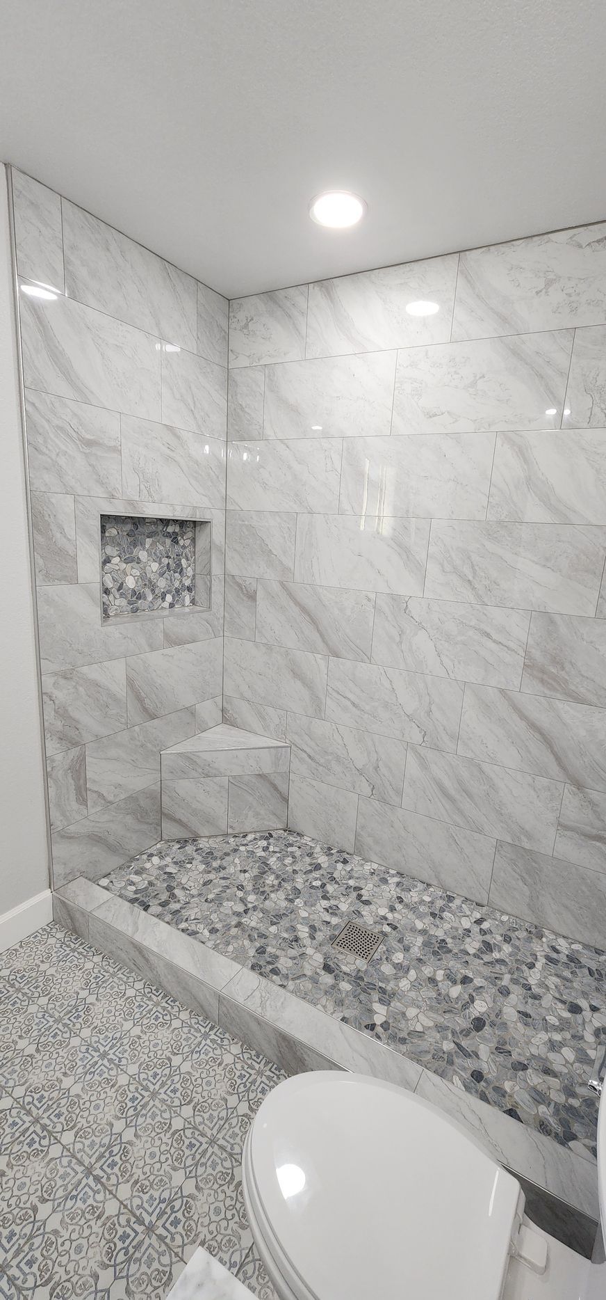 Modern gray-tile shower with a recessed shelf, pebble floor, and part of a toilet visible in the foreground