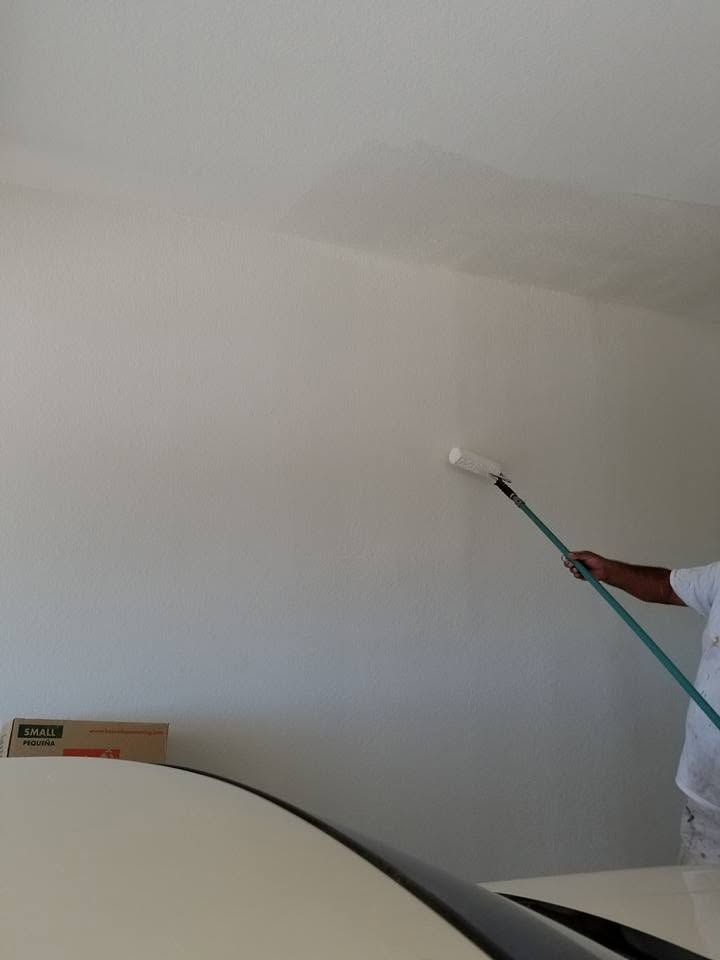 Person painting a white wall with a paint roller near a bed.
