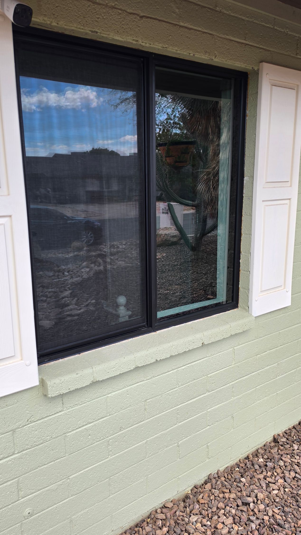 Black-framed window with white shutters on a light green house exterior, reflecting sky and trees.