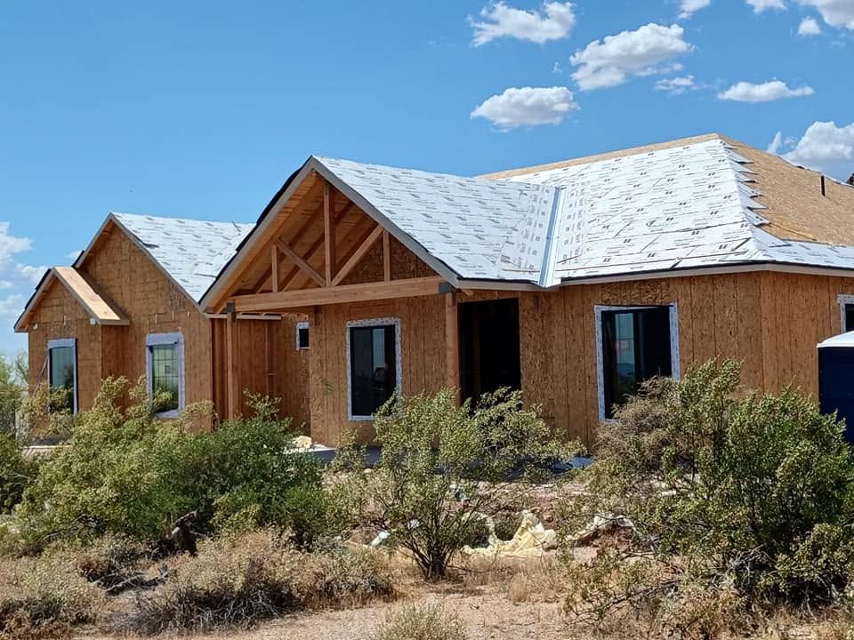 Partially built wooden house with white roof in a dry desert lot under a blue sky