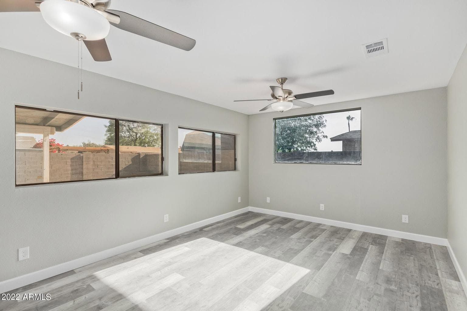 Empty light-gray living room with gray floors, ceiling fans, and large windows overlooking a fenced yard