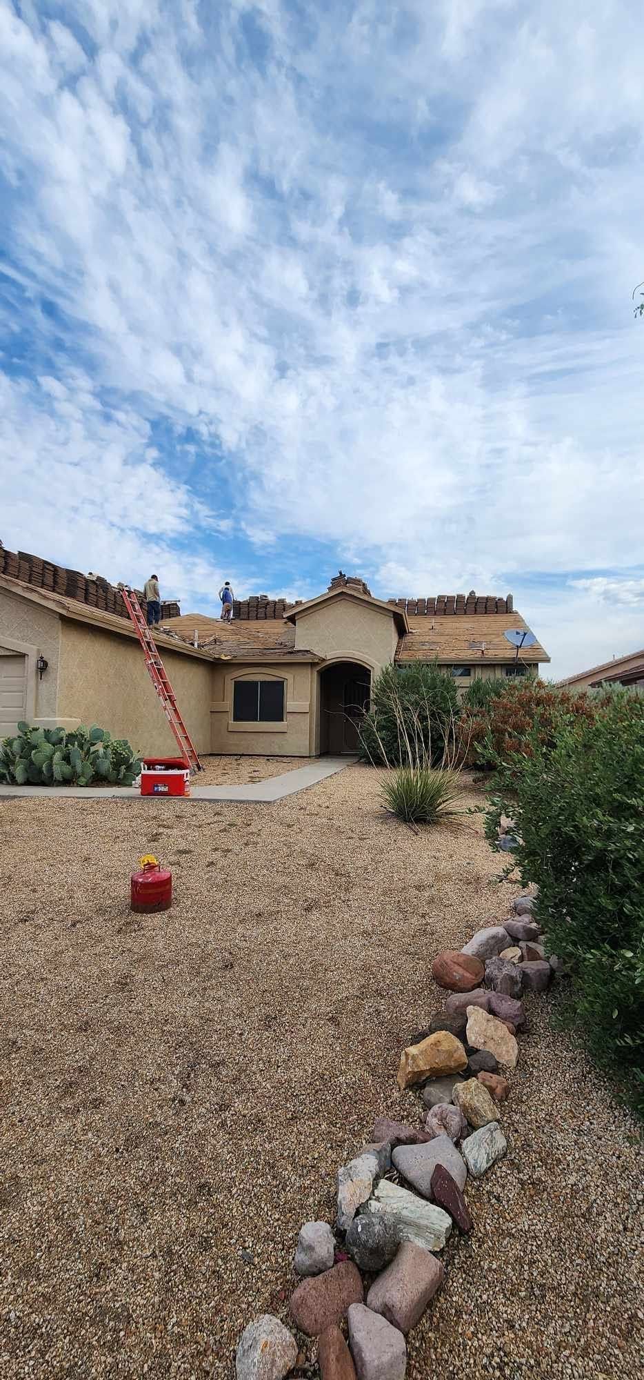 Stone courtyard leading to a rustic house under a blue, cloudy sky, with red planters and a rock border