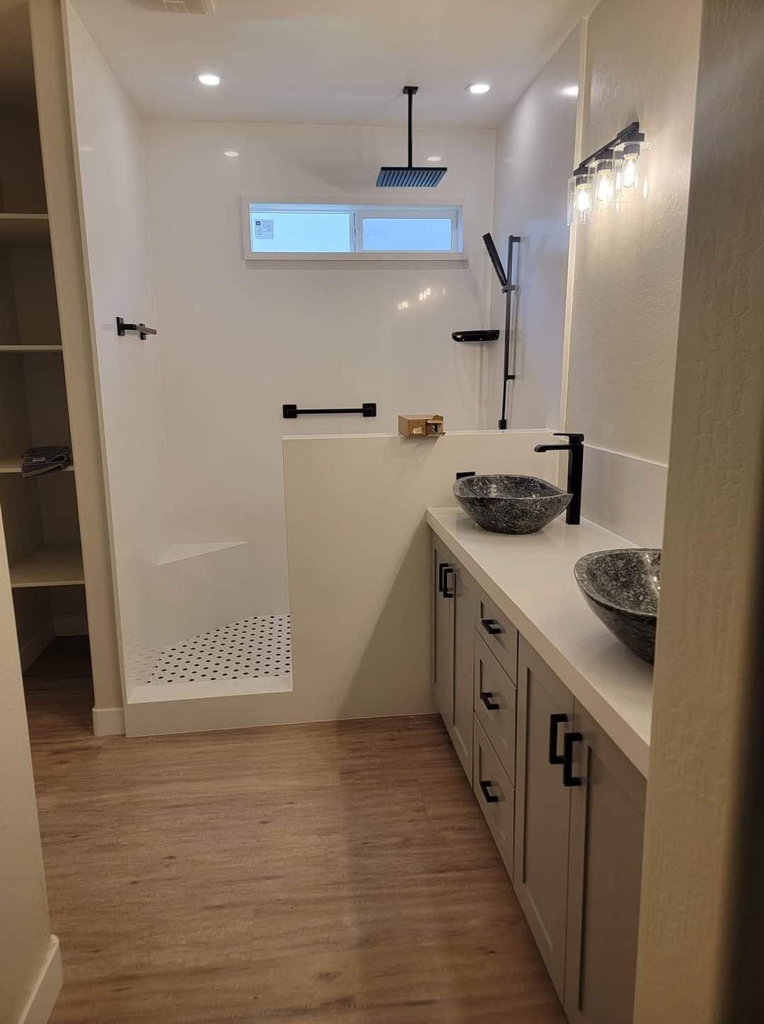 Modern bathroom with double sinks, wood floor, glass shower, and a small high window.