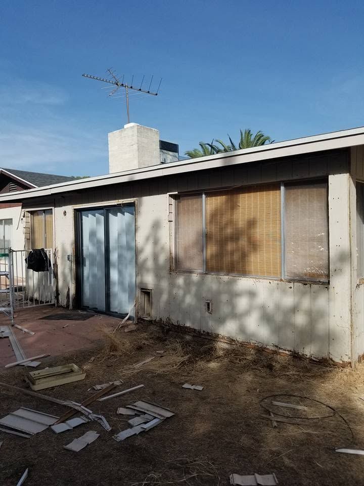 Single-story house exterior with white walls, blue door, shuttered windows, and a TV antenna on the roof.