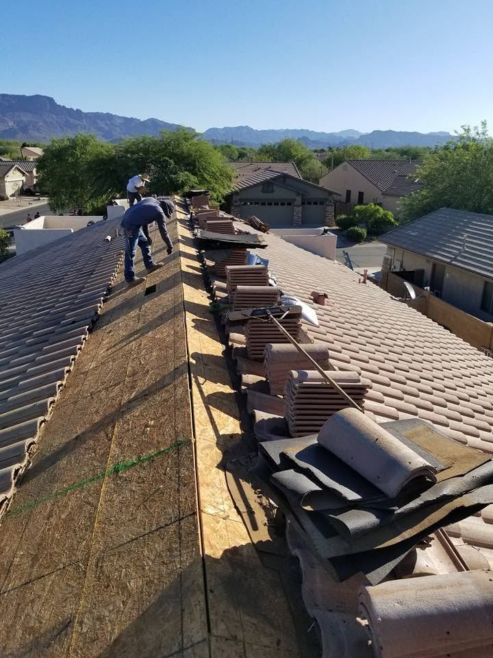 Worker on a rooftop removing shingles, with stacked materials and mountain homes in the background.