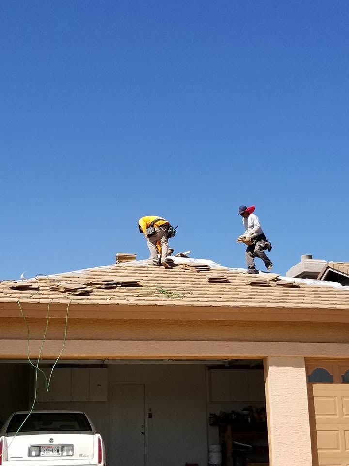 Two roofers working on a tan house roof, one kneeling and one standing under a clear blue sky