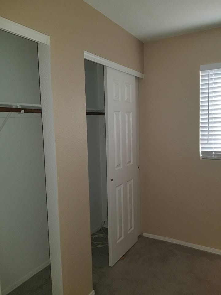 Empty bedroom closet with open sliding doors, beige walls, carpet, and a window with blinds.
