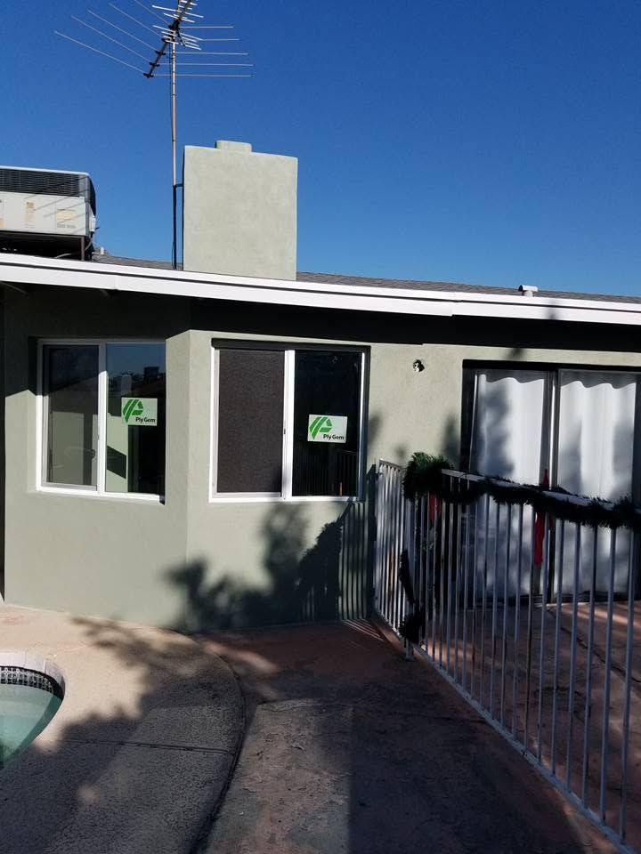 Single-story house with green doors, white curtains, and a fenced front walkway under a blue sky