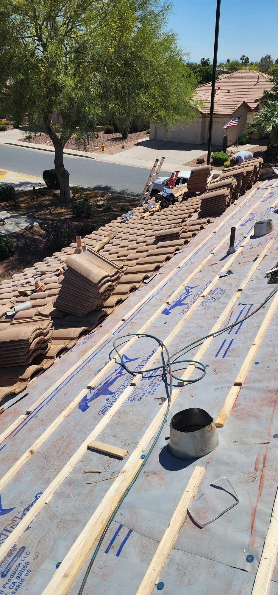 Roof under construction with stacked shingles, tools, and a tree-lined street in the background