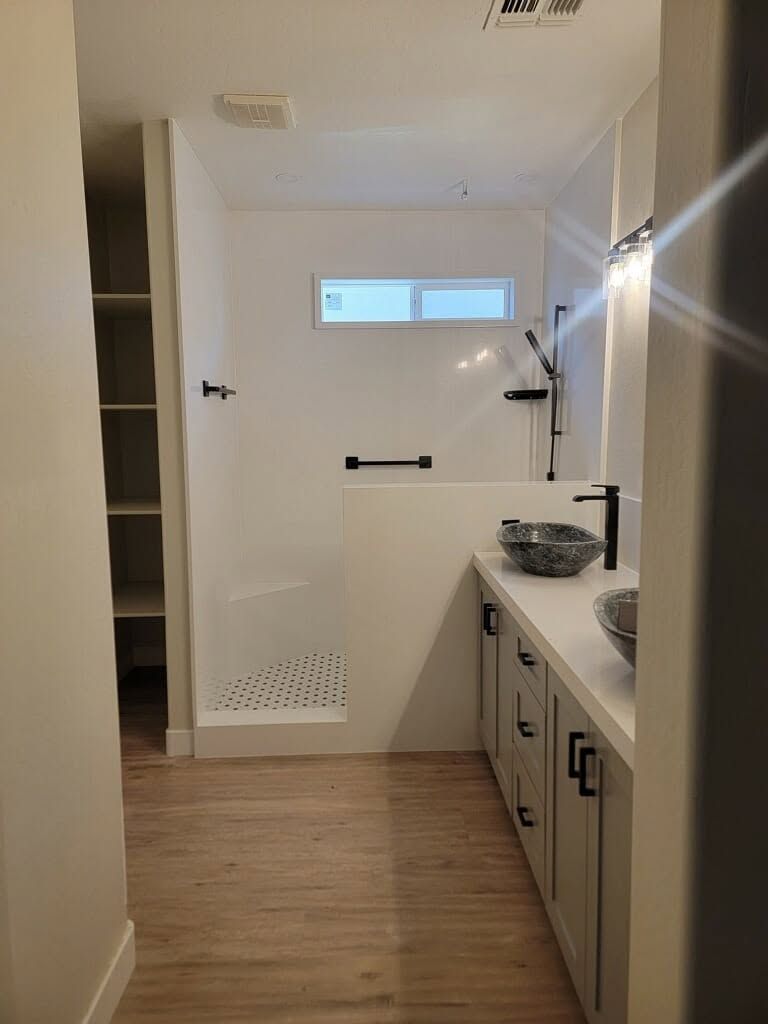 Bright bathroom with white walls, wood floor, shower area, sink counter, and small window.