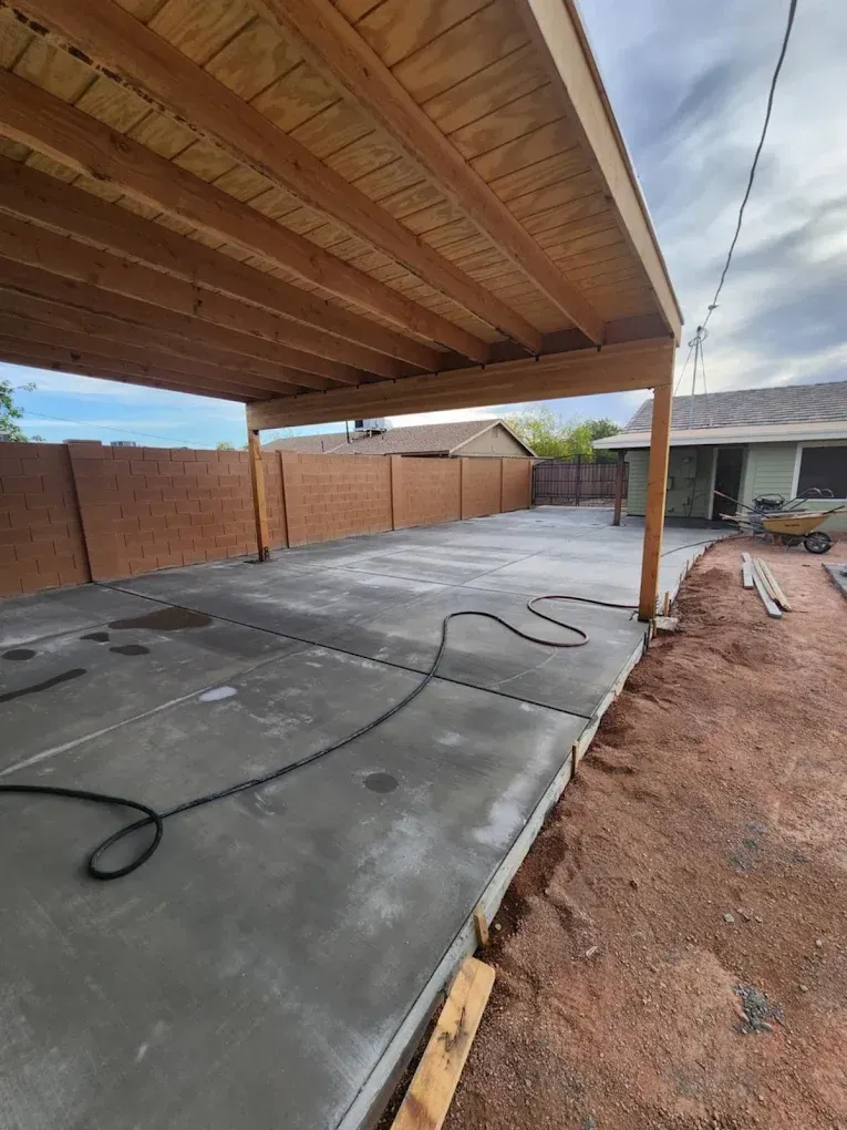 Concrete patio under a wooden pergola beside a brick wall, with hoses on the ground and a cloudy sky.
