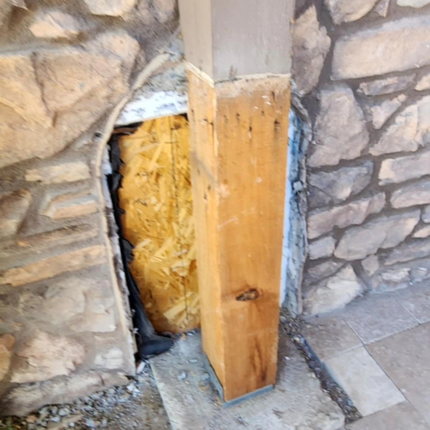 Damaged stone wall with exposed insulation and a vertical wooden post at the corner