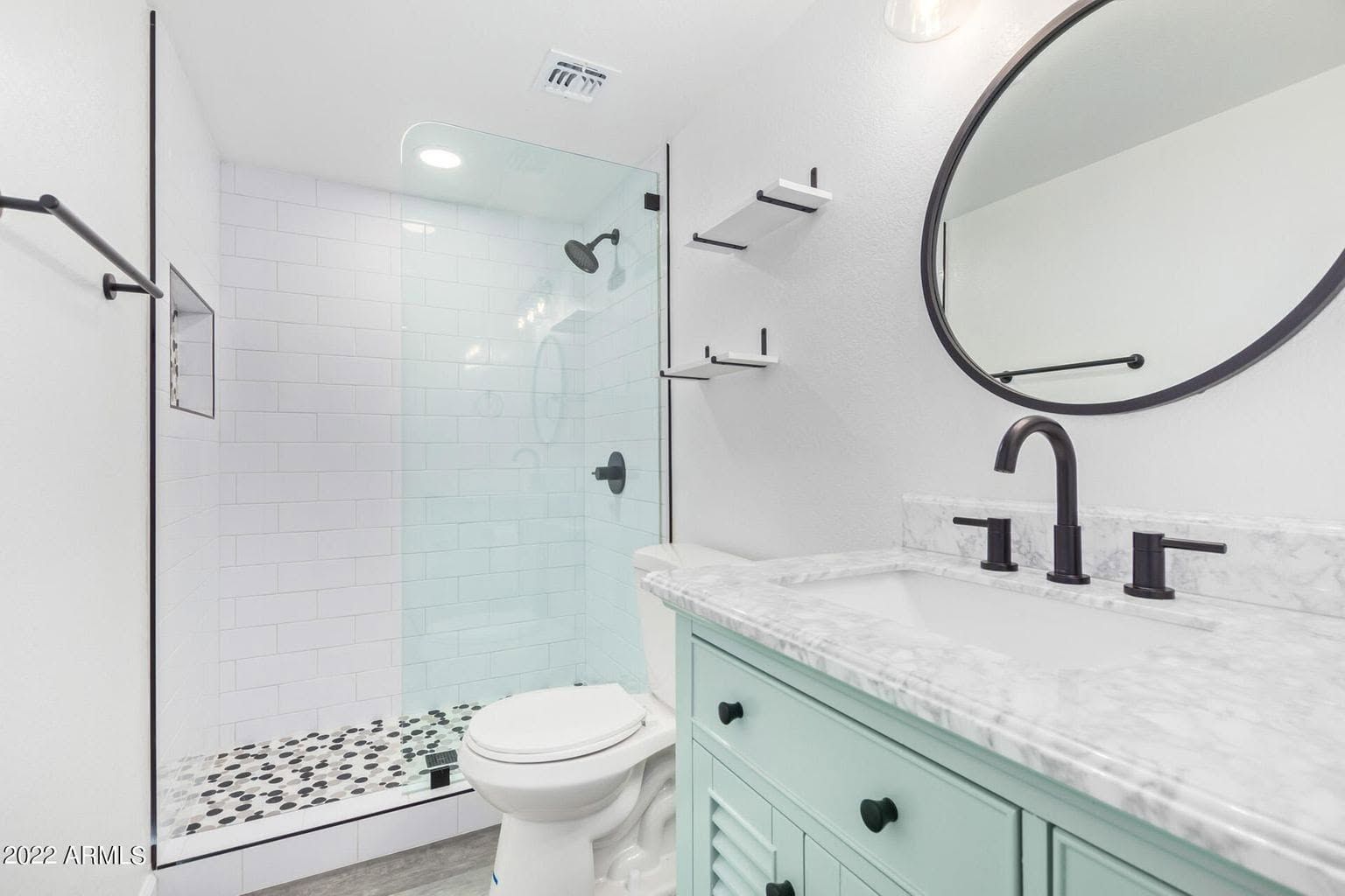 Bright modern bathroom with mint vanity, black fixtures, round mirror, toilet, and glass shower.