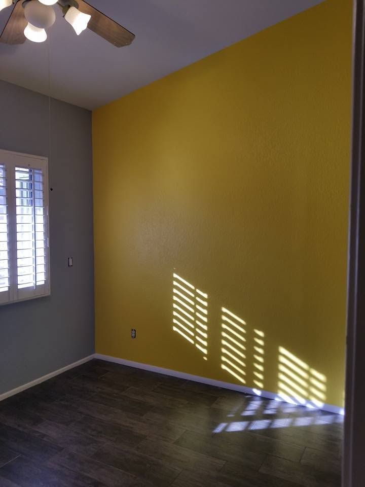 Empty room with yellow accent wall, gray walls, dark wood floor, and sunlight through blinds.