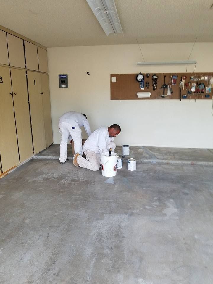 Two workers in white uniforms painting a garage floor with buckets and tools nearby