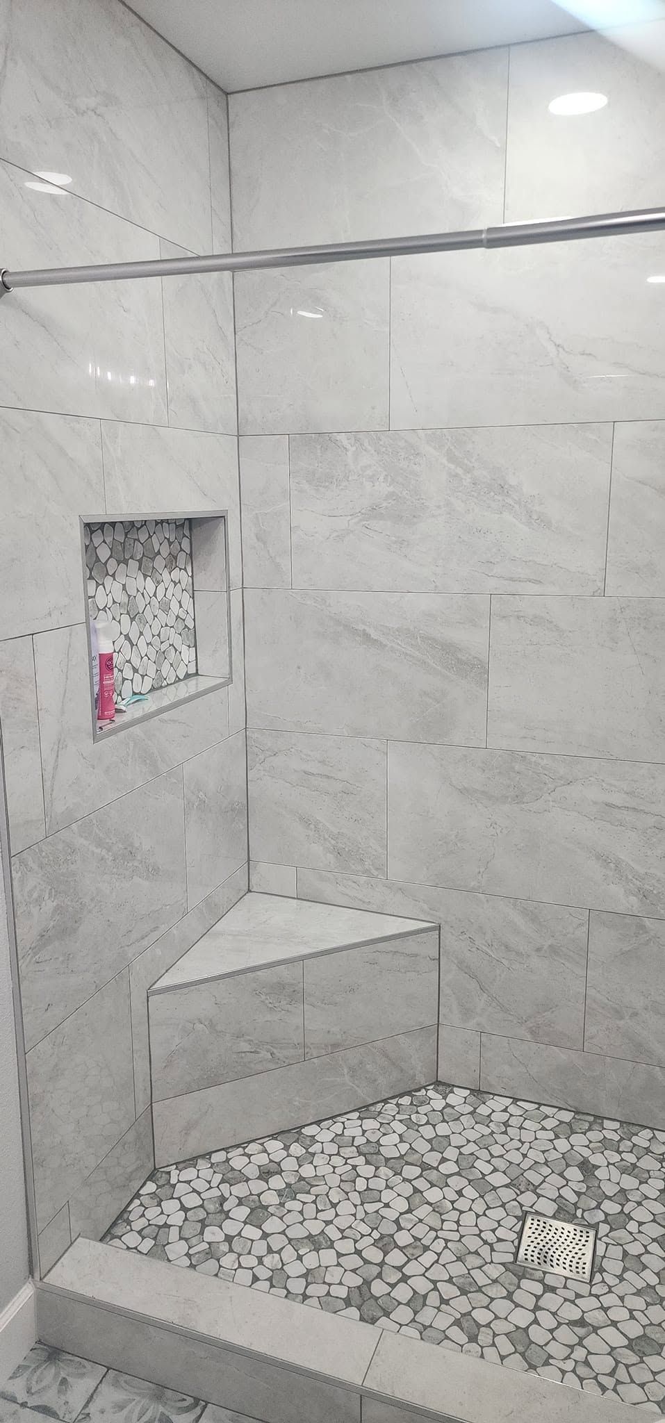 Small tiled shower with a built-in corner shelf and pebble-pattern floor.