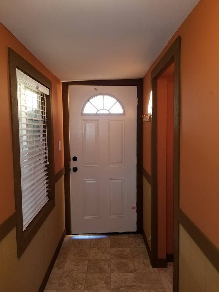 Narrow hallway with tile floor leading to a white front door, flanked by windows and wood trim.