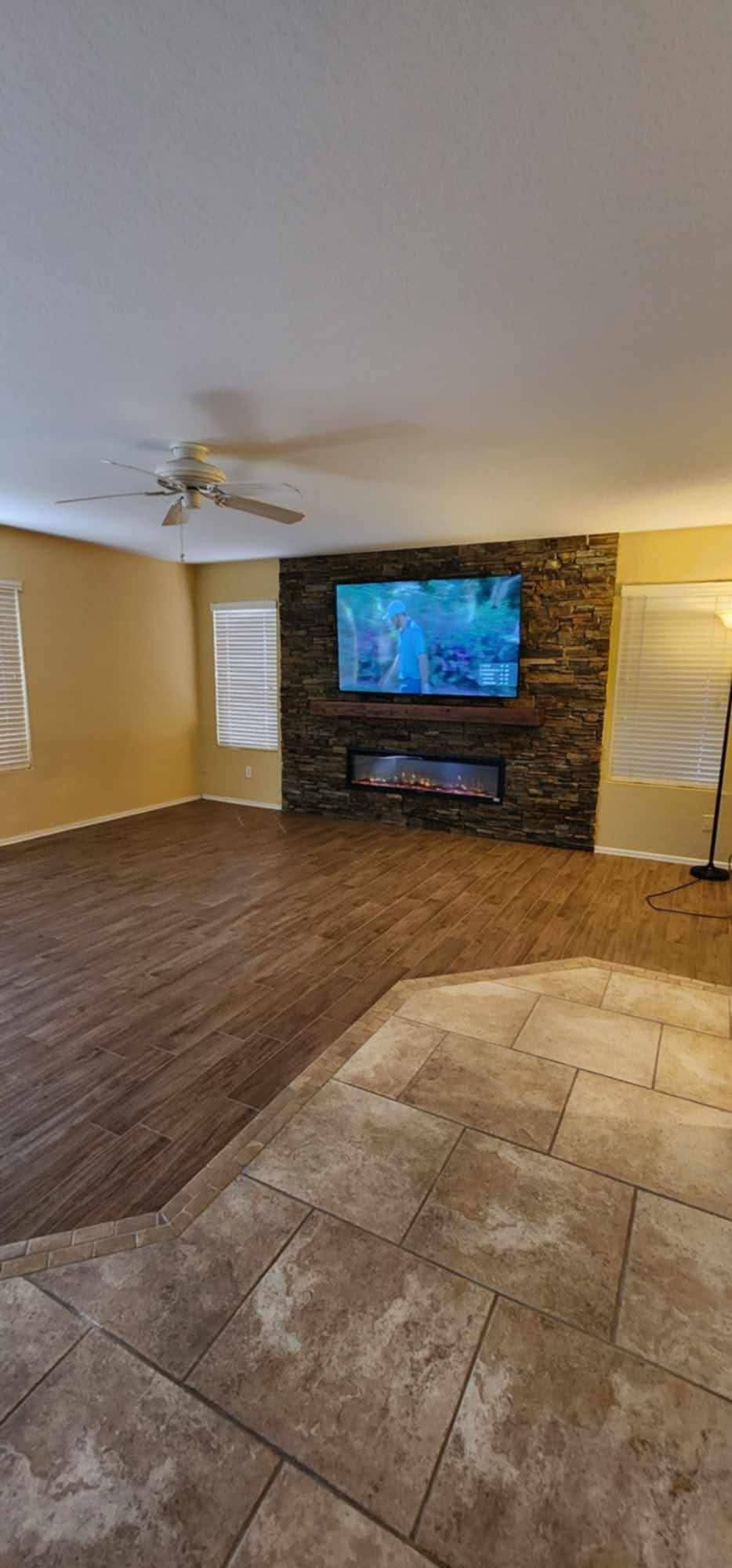 Empty living room with wood and tile flooring, ceiling fan, stone fireplace, and wall-mounted TV.