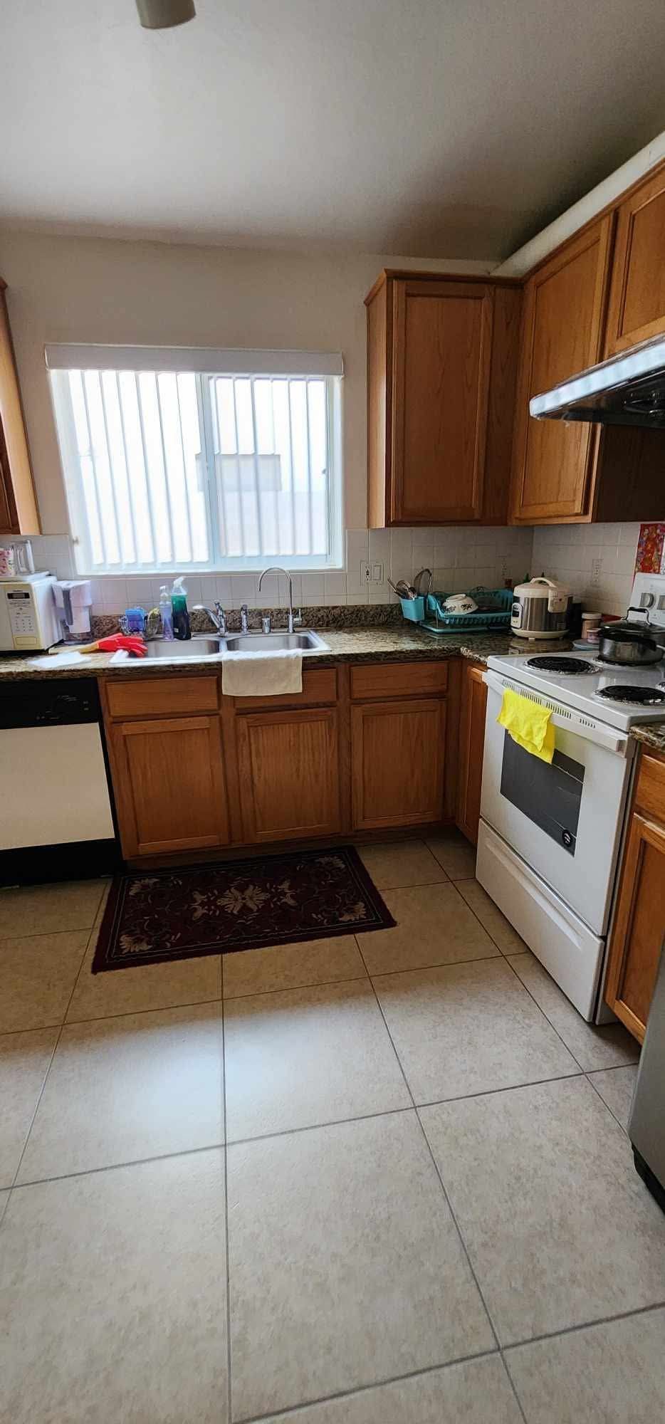 Small kitchen with wooden cabinets, white tile floor, sink under a window, and stove with yellow towel.
