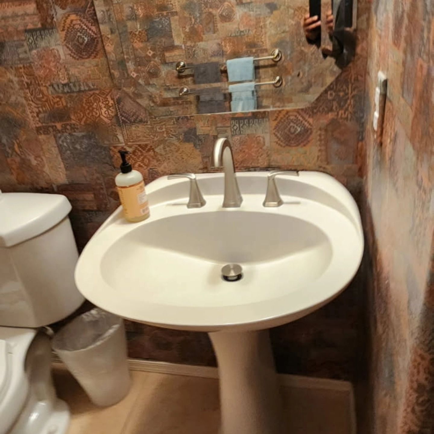 Small bathroom sink with soap dispenser, mirror, towel rack, and toilet beside a patterned tiled wall.