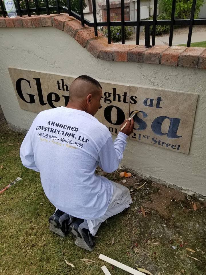 Man kneeling and painting a stone sign along a sidewalk in a grassy area