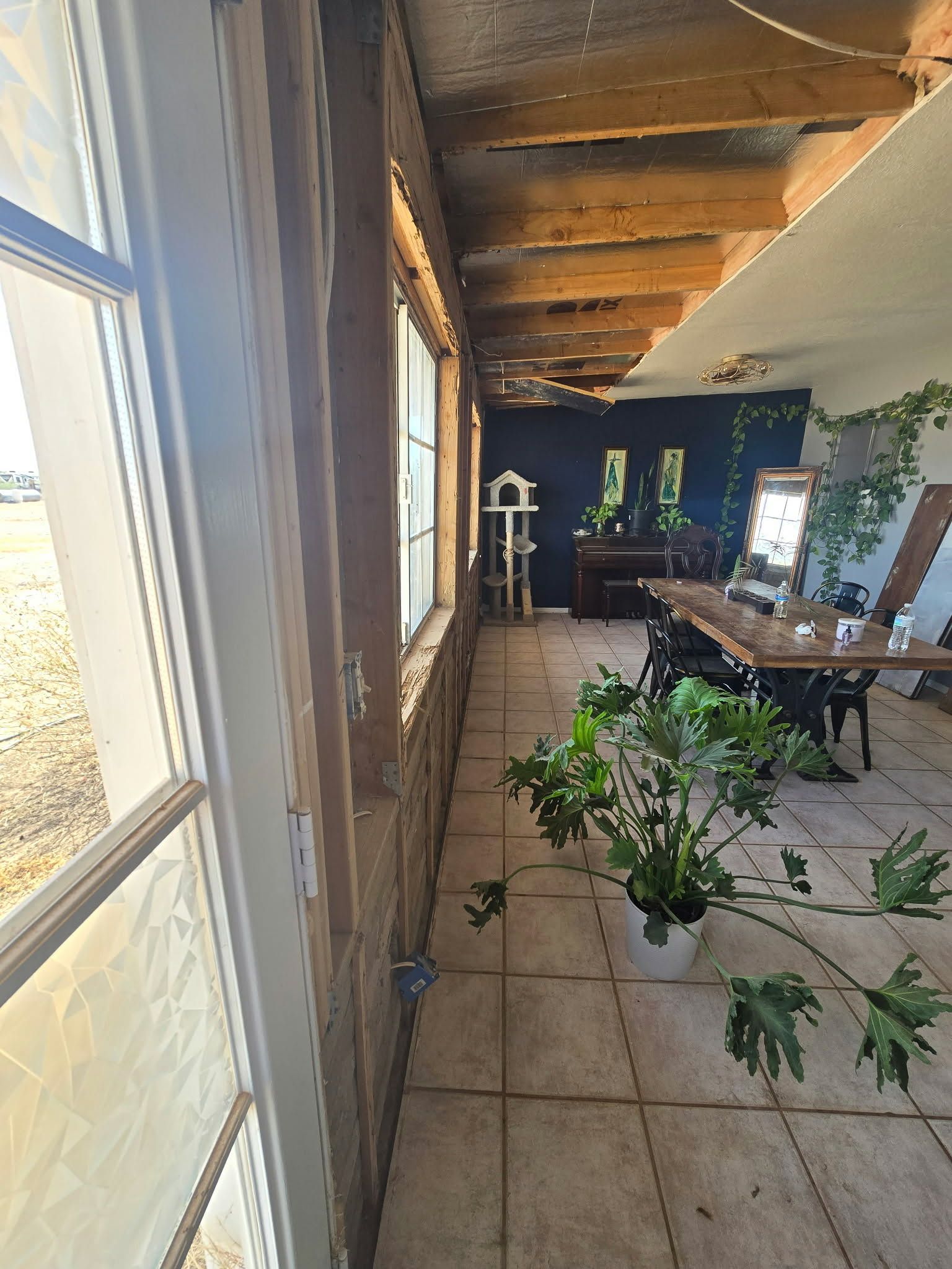 Narrow covered patio with tile floor, wooden ceiling, table, chairs, and potted plants by a window.