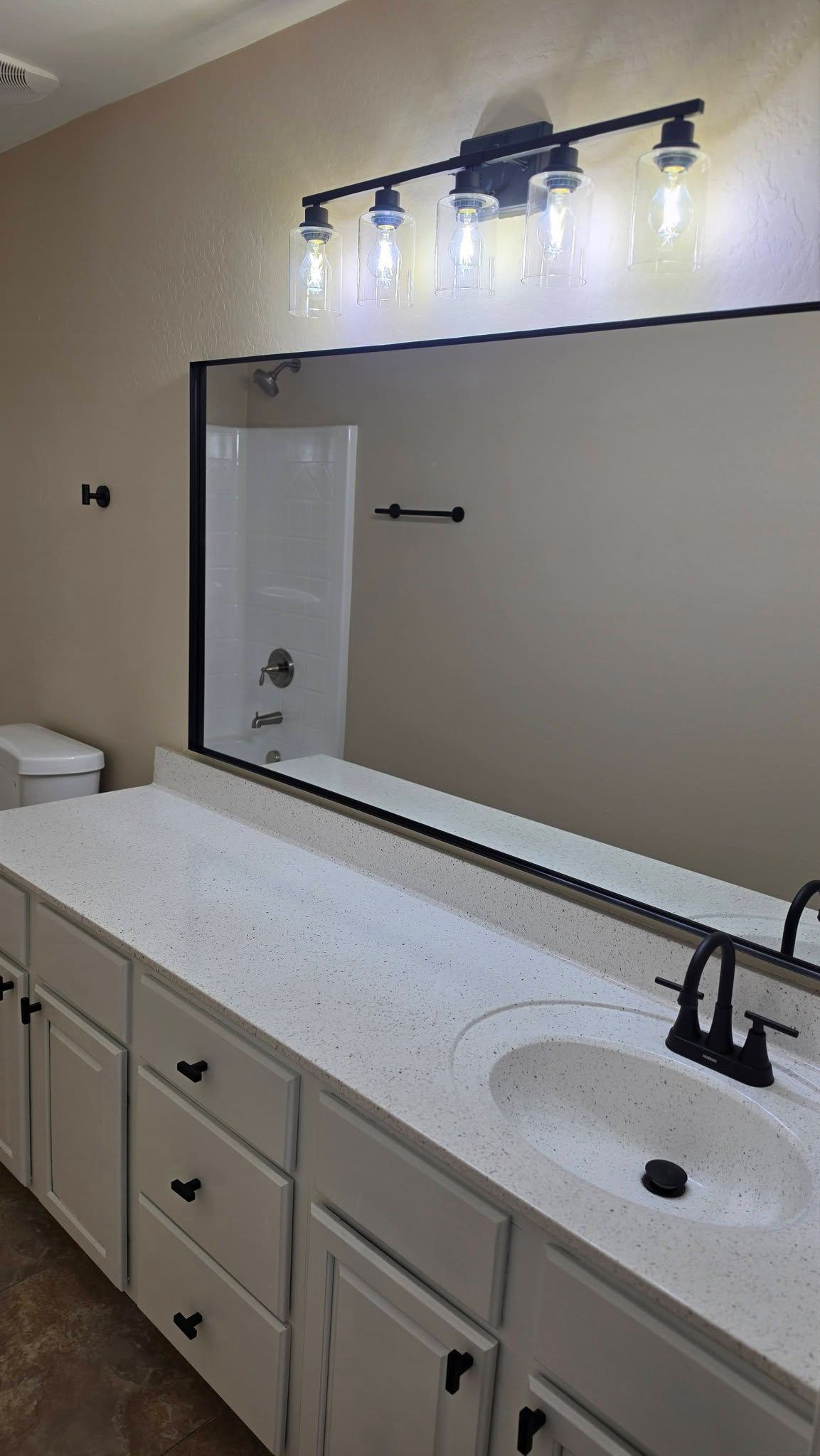 Bathroom vanity with large mirror, lit wall sconce, white countertop, and black fixtures.