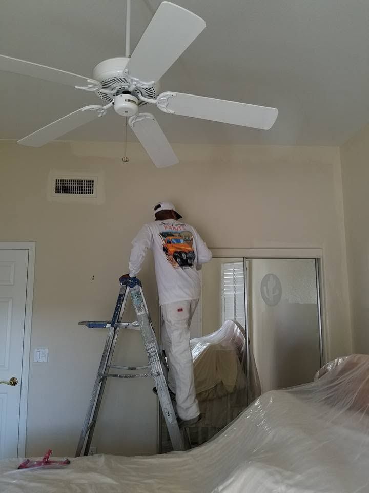 Painter on a ladder painting a bedroom ceiling fan in a room with covered furniture.