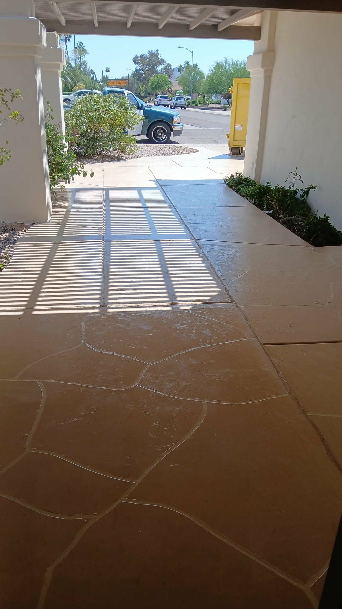 Covered walkway with sunlit tiled floor leading to a parking lot and blue vehicle beyond.