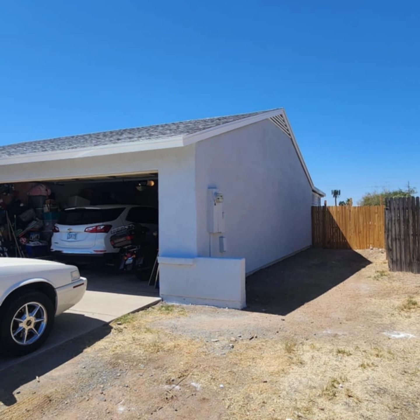 Single-story garage with open door, parked cars, and a wooden fence on a dry dirt lot under a blue sky