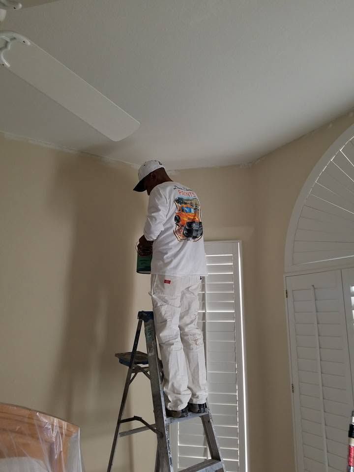 Person painting a beige ceiling while standing on a ladder in a room with white plantation shutters.