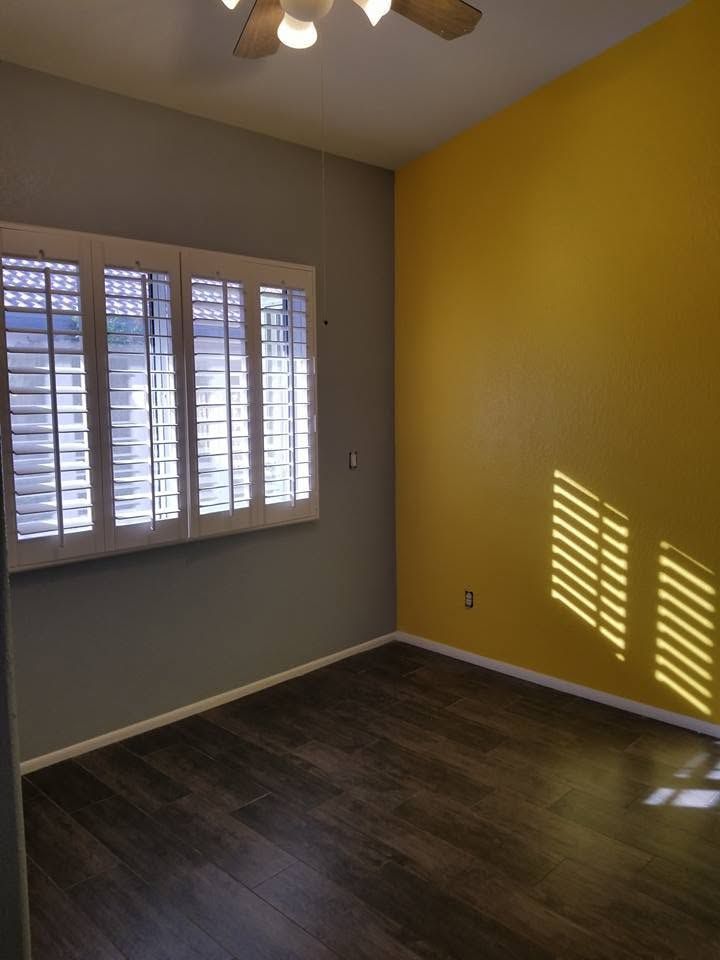 Empty room with dark wood floor, gray and yellow walls, and sunlight streaming through window blinds.