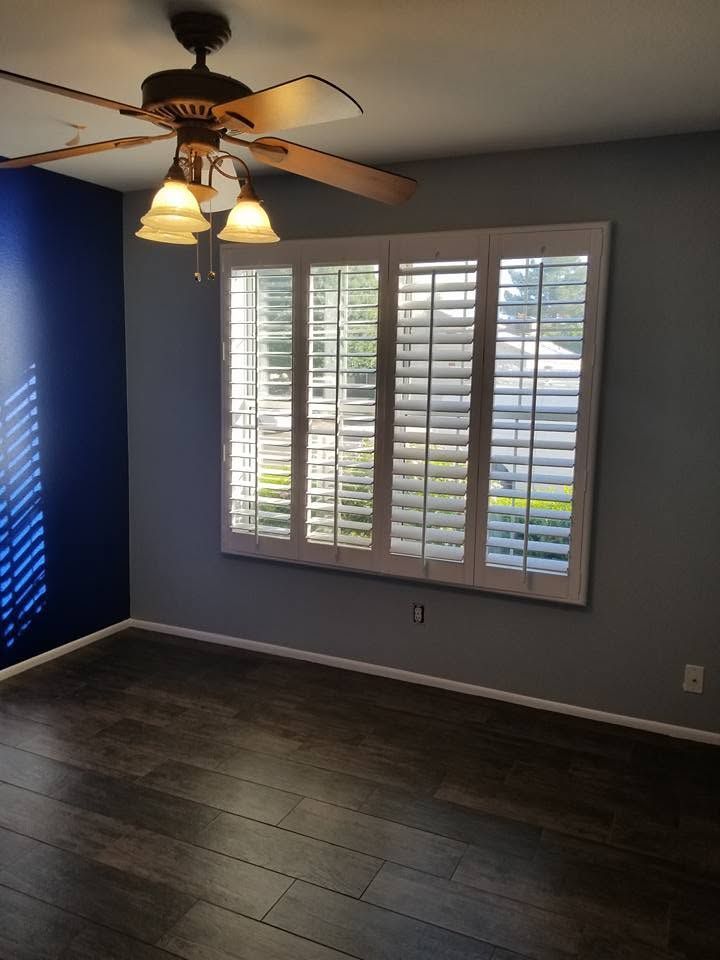 Empty room with dark wood floors, gray walls, window shutters, and a ceiling fan with lights.