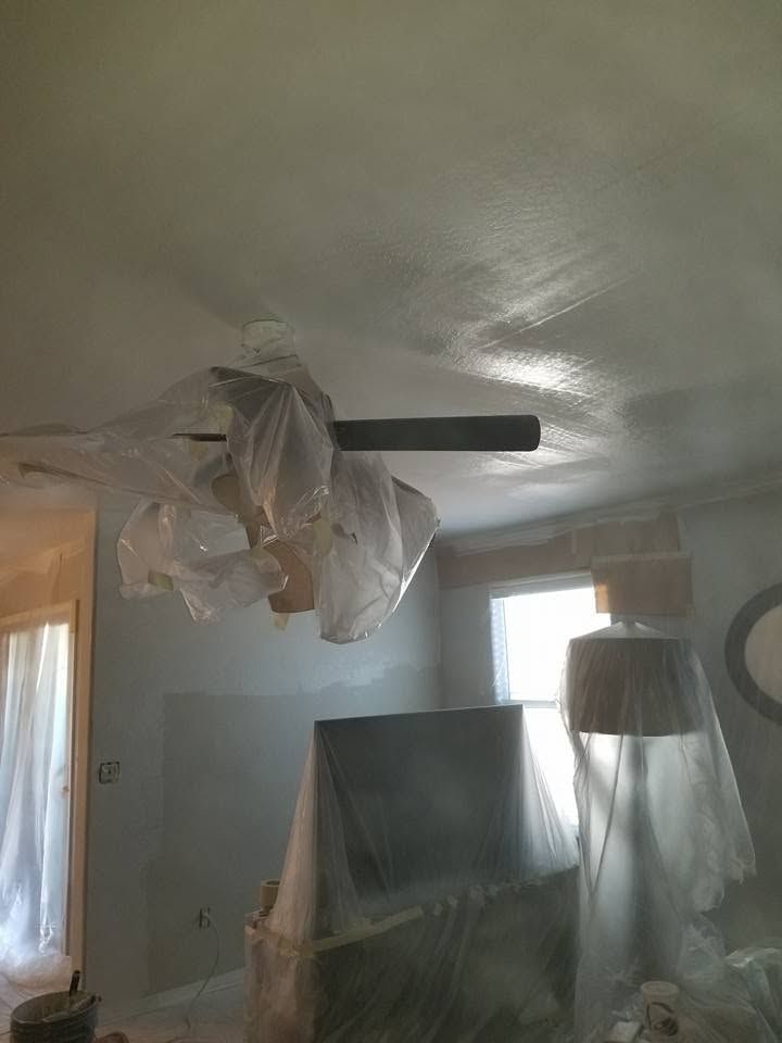 Plastic-covered furniture in a room under renovation with a ceiling fan and curtained windows.
