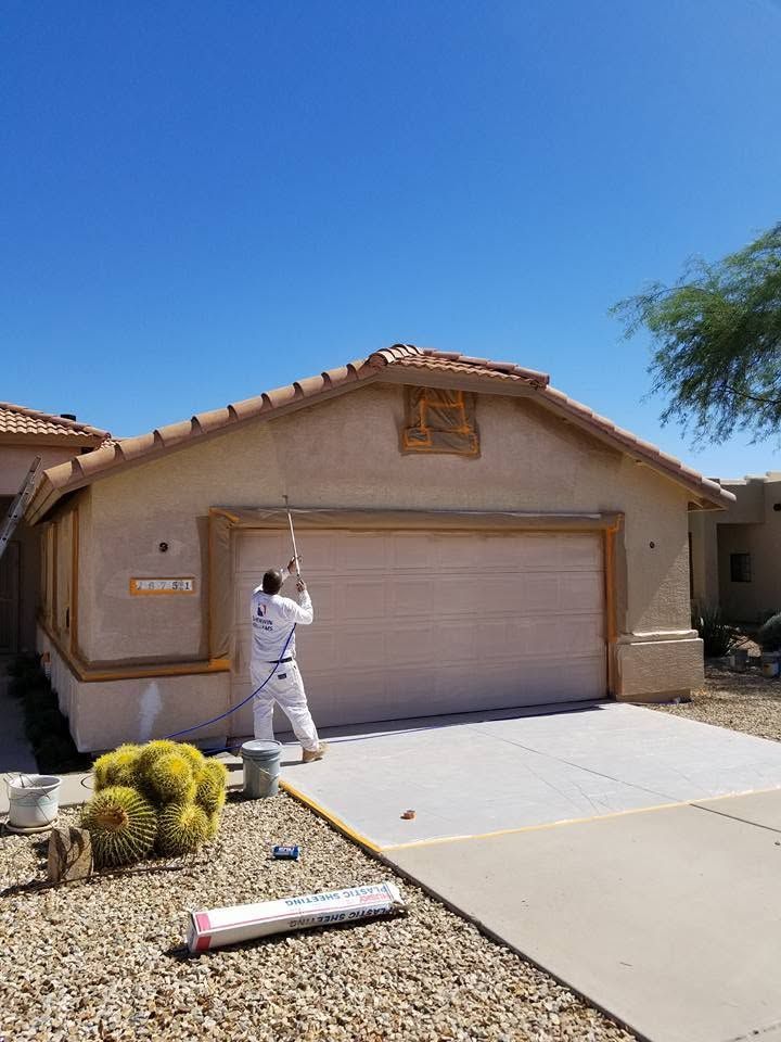 Worker painting a beige stucco house exterior and garage in bright sunlight
