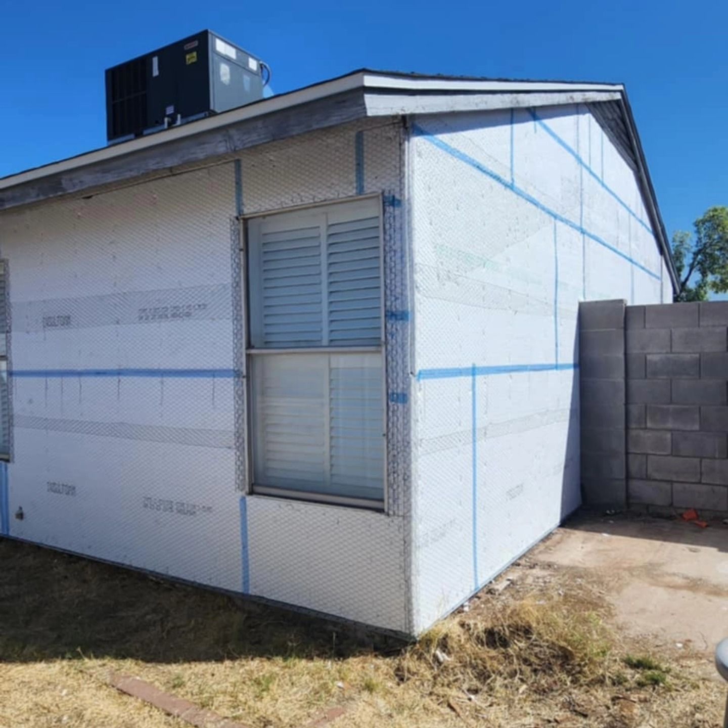Small white house exterior with blue trim, window shutters, and a cinder block wall beside it.
