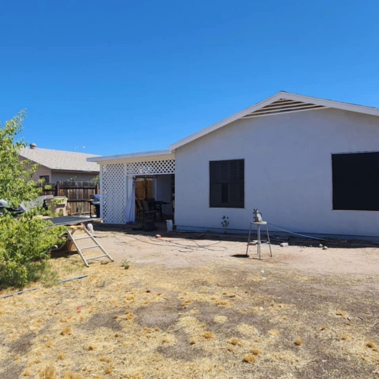Single-story house exterior with white walls, black windows, and a dry front yard under a clear blue sky