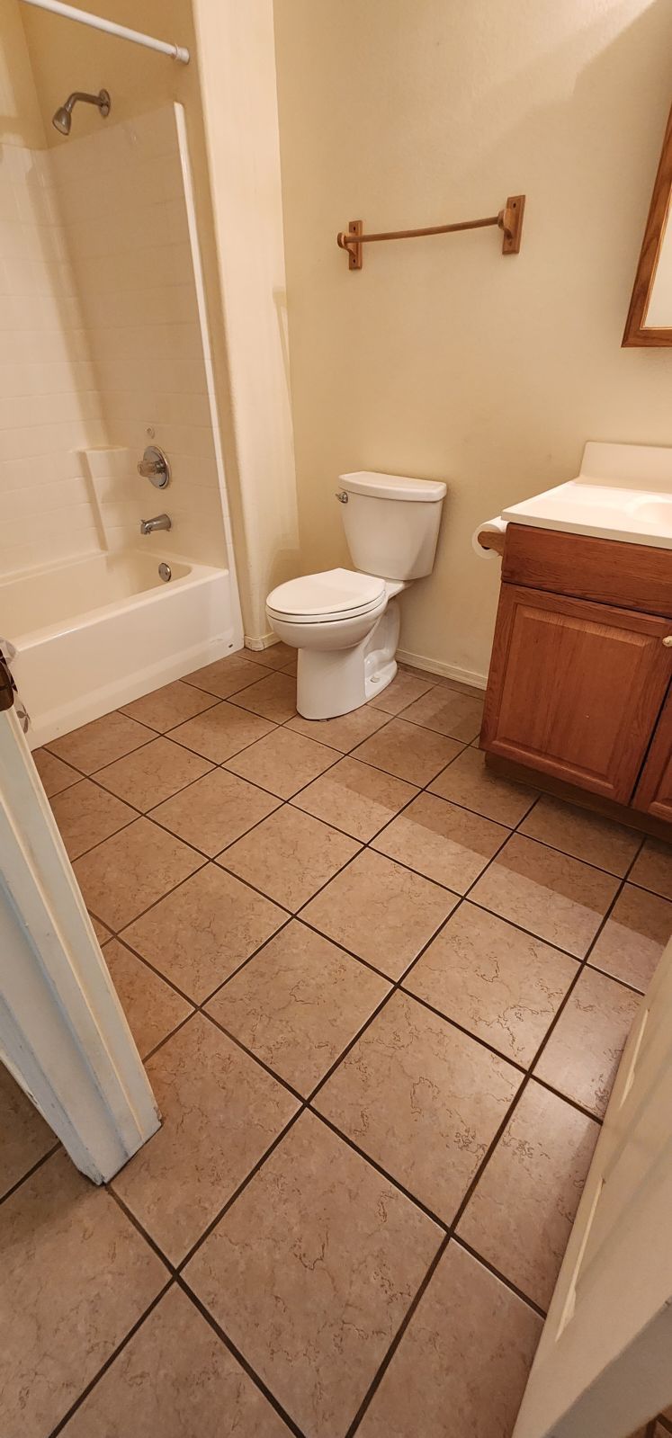 Small beige bathroom with a toilet, tub, wood vanity, and tiled floor.