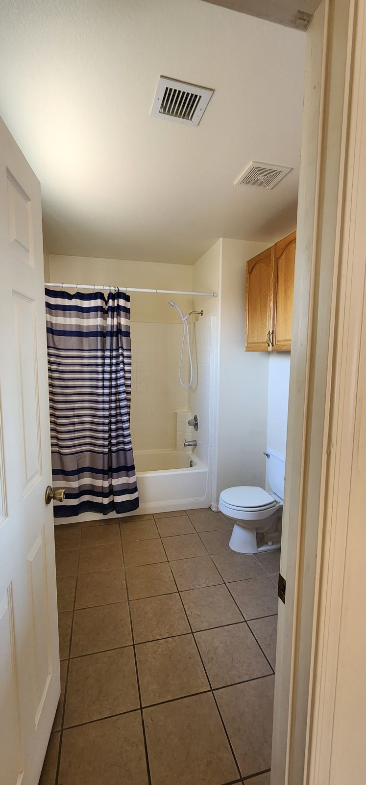 Small bathroom with tan tile, striped shower curtain, white toilet, and wooden cabinet.