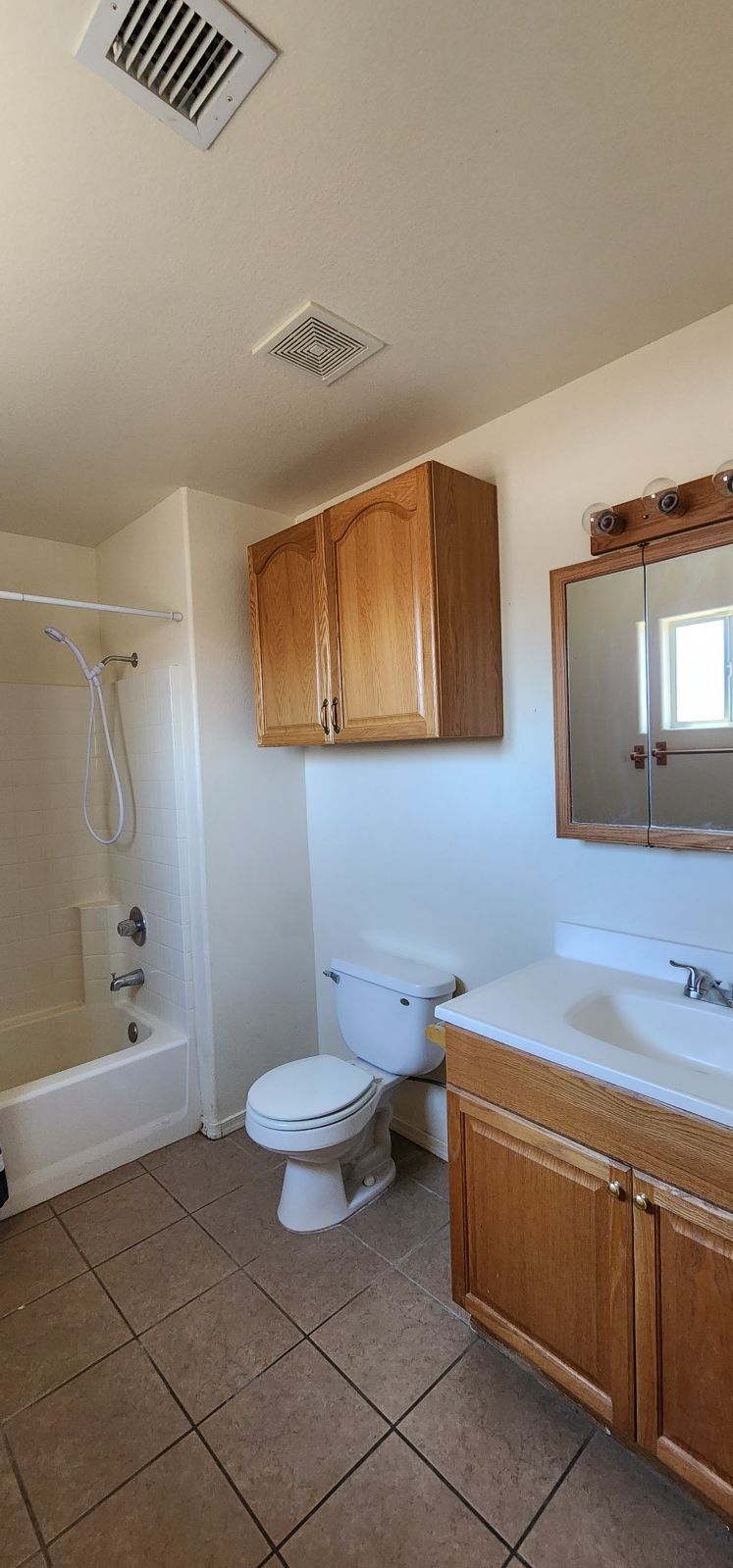 Small bathroom with white toilet, sink, bathtub, wood cabinets, and tan tile floor.