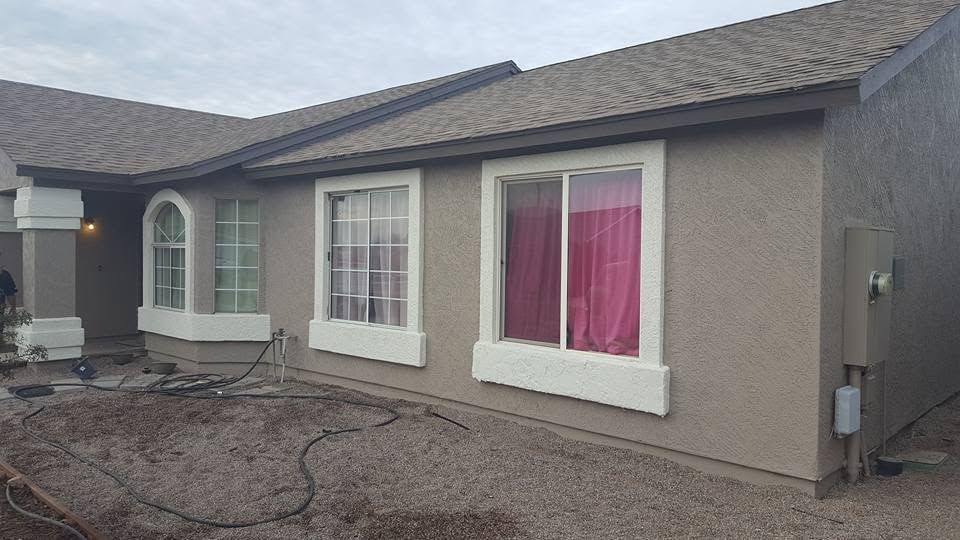 Single-story stucco house exterior with white-trimmed windows and pink curtains, gravel yard in front.