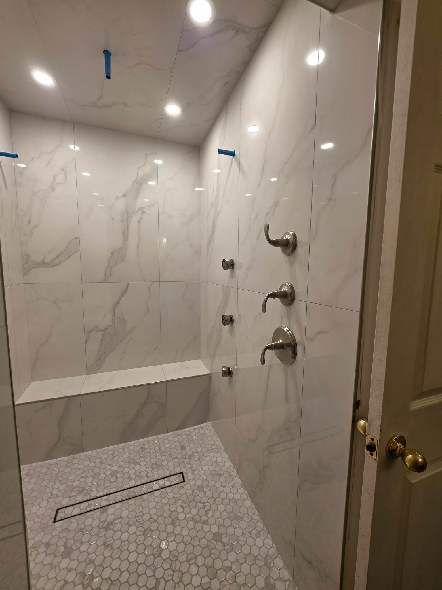 Modern tiled walk-in shower with bench, mosaic floor, and wall-mounted fixtures, viewed through an open door