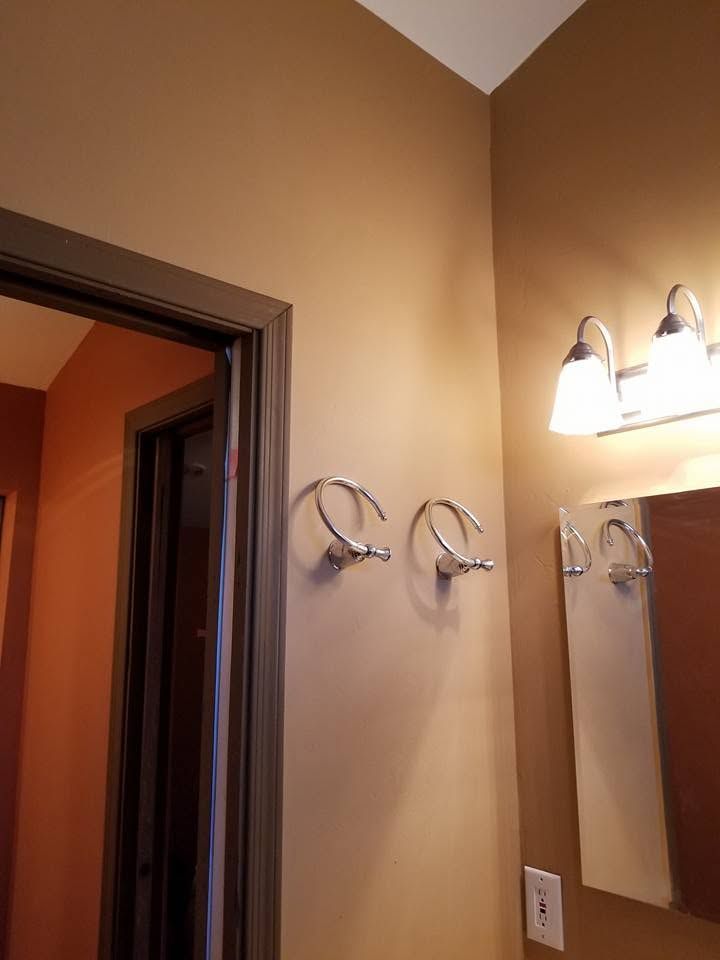 Bathroom corner with beige walls, mirror, and lit vanity light above a towel rack.