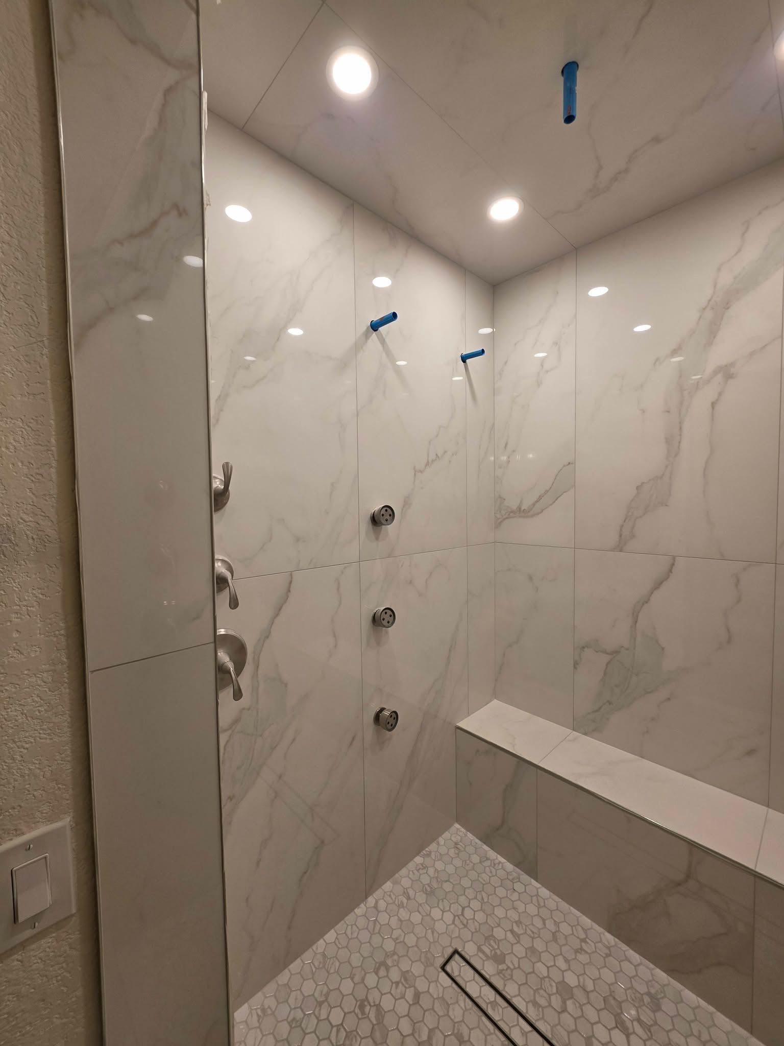 Modern tiled shower under construction with marble-look walls, ceiling lights, and blue tape on fixtures